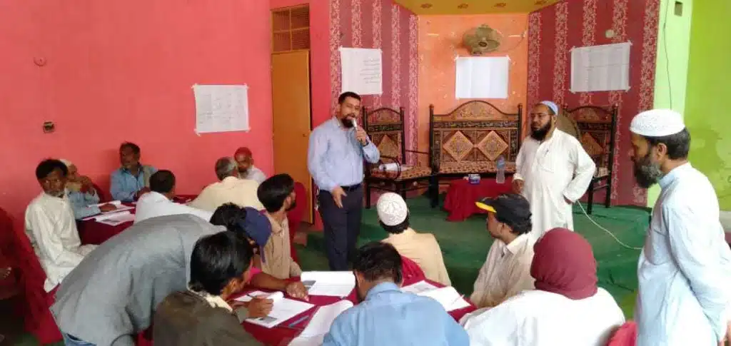 A community workshop in a hall, with participants seated at low tables and a facilitator standing at the front.