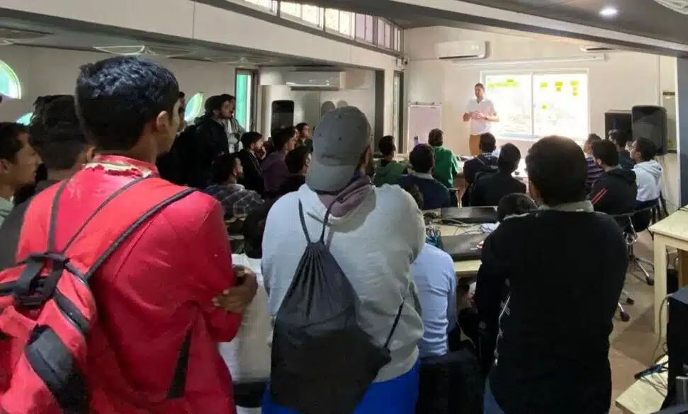 A conference audience standing and seated in a hall, facing a presentation.