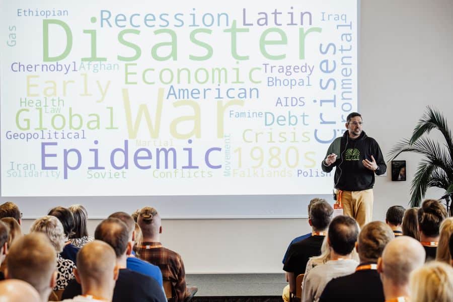 Kubair Shirazee speaking at Agile Saturday Tallinn 2024 in front of a word cloud of crises such as war and recession